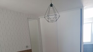 Una camera da letto con un letto, una lampada a sospensione geometrica, una parete bianca decorata con motivi e una porta che dà su un'altra stanza.