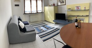 En stue med sofa, fjernsyn, træbord og et køkkenområde med skab og hylde.