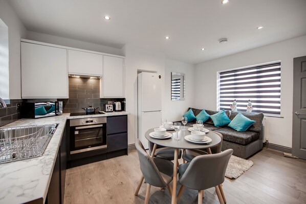 A modern kitchen with white cabinets, a dining area with a round table, and a grey sofa with blue cushions. - 4 Bed - Sleeps 6 - Private Garden - Free Parking (Ashford)
