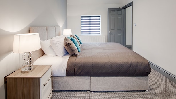 A modern bedroom with a bed, bedside table, lamp, and a window with blinds.
