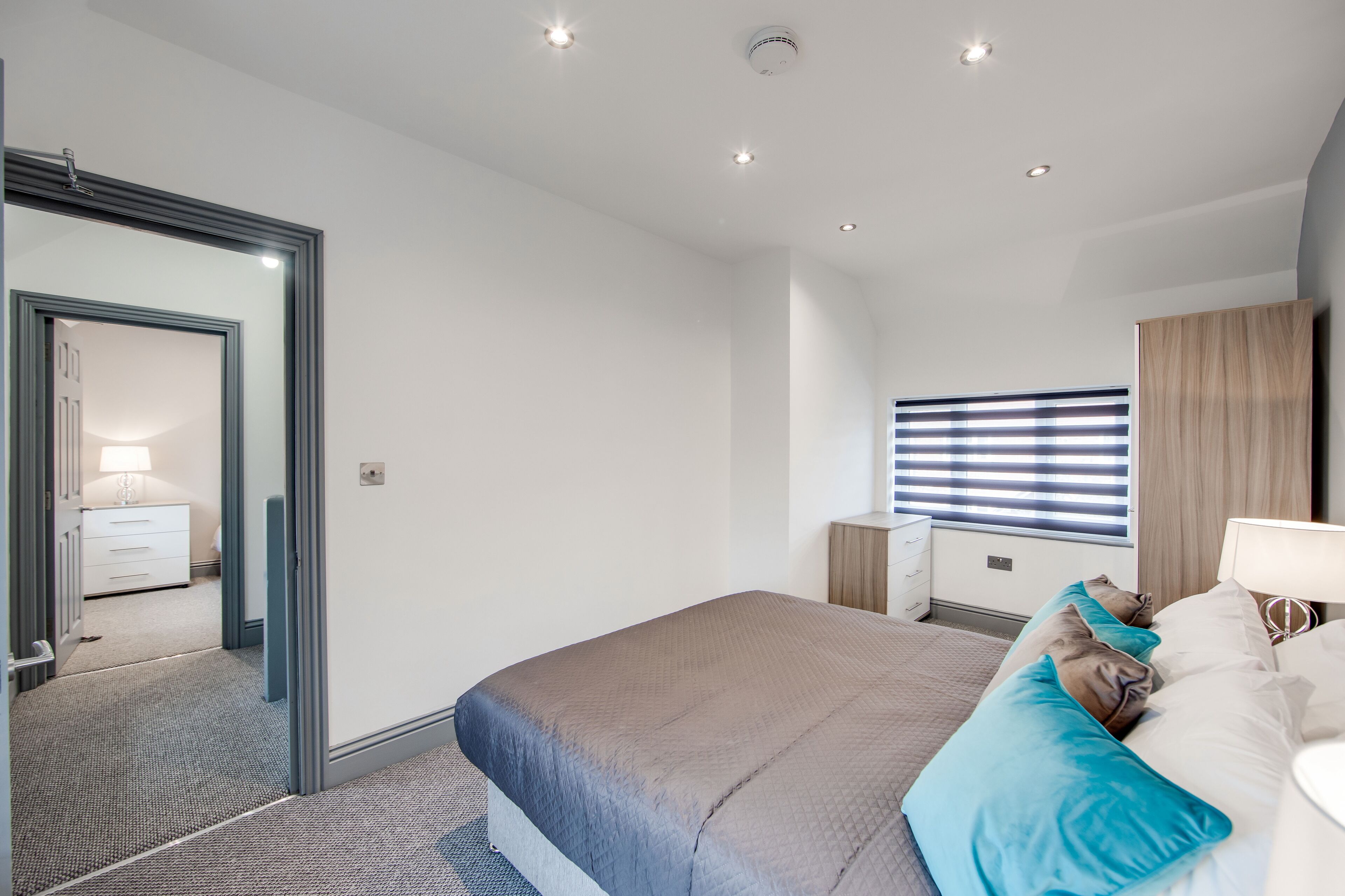 A modern bedroom with a large bed, a bedside table, a mirror, and a window with blinds.