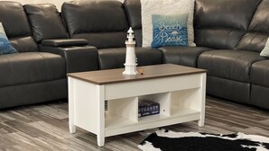 Una mesita de noche de madera blanca con un modelo de faro, un tapete de cuero y un sofá de cuero con un cojín que dice 'Beach Please'.