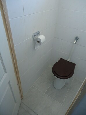 Um banheiro pequeno e limpo, com azulejos brancos, vaso sanitário com assento marrom e rolo de papel higiênico na parede.