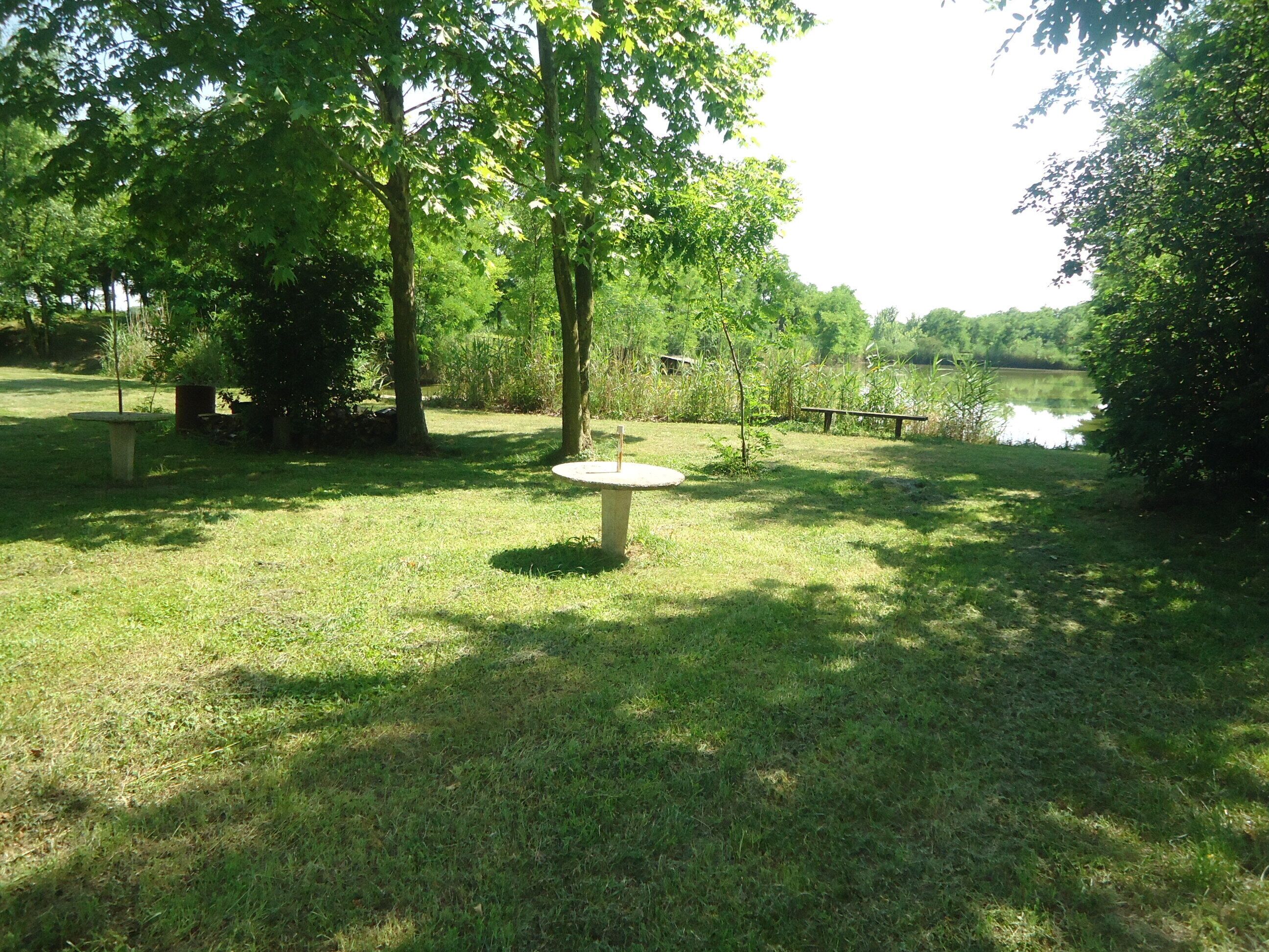 Um parque com uma mesa de pedra circular, grama, árvores e um corpo de água.