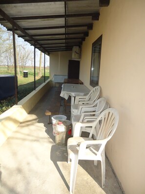 Uma área de pátio externo com mesa, cadeiras, um contentor de lixo e uma janela.