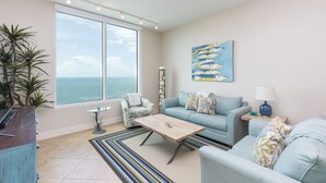 A living room with a large window, a sofa, a coffee table, and a painting of fish. - Breathtaking Views from 23rd Floor at Sapphire! Resort Pool, Hot Tub, Spa, Movie Theater & More! (South Padre Island)