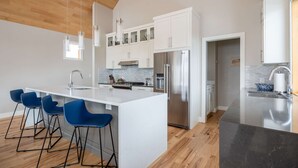 Eine moderne Küche mit weißen Schränken, einem Edelstahlkühlschrank und einer Marmarbeitsplatte. Drei blaue Barhocker und eine Hängeleuchte sind vorhanden.