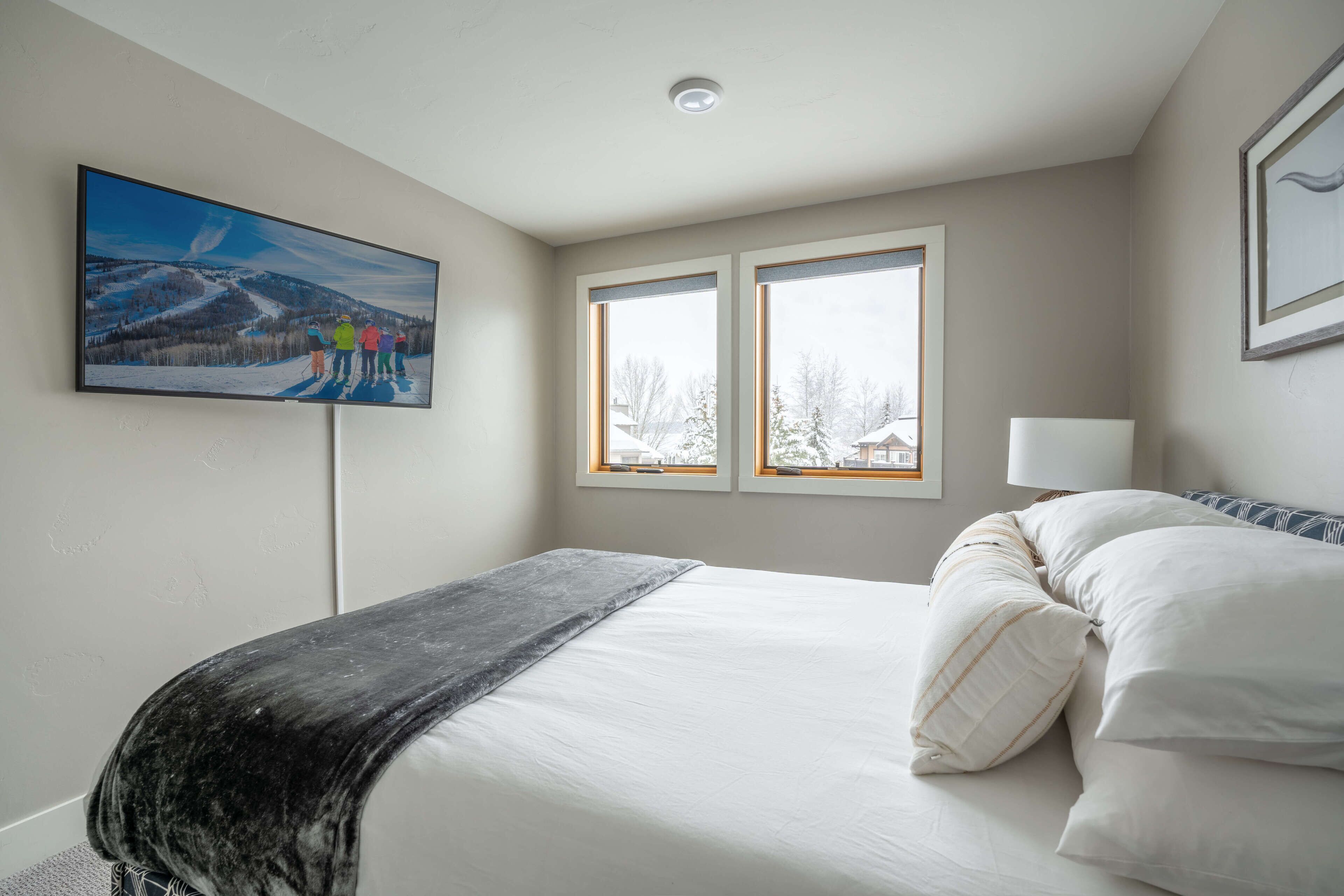 A bedroom with a bed, two windows, a painting, and a ceiling light.