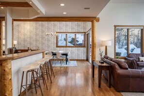 Une cuisine moderne avec un bar en bois, des tabourets de bar et un coin repas comprenant une table et des chaises. Une grande fenêtre offre une vue sur des arbres couverts de neige.