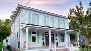 Une maison blanche à deux étages, avec des volets verts, un porche à l’avant et un perron en briques.