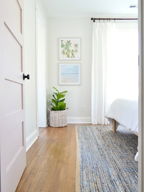 A bedroom with a bed, a window with white curtains, a wall-mounted picture, and a wooden floor. - Doublemint Townhouse, Walk to Beach & Downtown (Cape Charles)