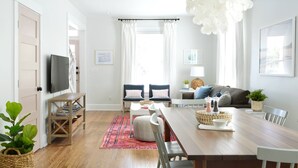 Un salon moderne avec une table à manger en bois, deux chaises, un canapé, un téléviseur fixé au mur et un grand lustre.