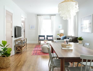 A modern living room with a wooden dining table, two chairs, a sofa, a TV on the wall, and a large chandelier. - Doublemint Townhouse, Walk to Beach & Downtown (Cape Charles)