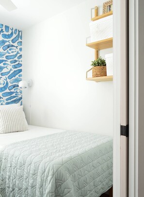 Une chambre avec un lit, un mur orné d’un motif représentant une baleine bleue, une lampe blanche et des étagères avec des plantes.