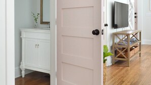 Une pièce avec un plancher en bois, une porte blanche, une coiffeuse blanche avec un miroir et un téléviseur posé sur un meuble en bois.