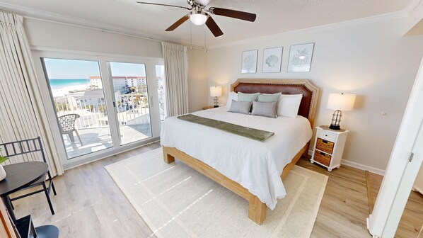 Une chambre d’hôtel avec un grand lit, un ventilateur de plafond, une table de chevet avec une lampe et une vue sur la plage depuis les portes du balcon.