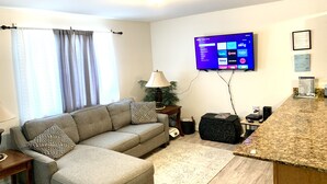 Ein Wohnzimmer mit einer Couch, einem flachbildfernseher an der Wand, einer Lampe und einem kleinen Tisch mit einem Korb.