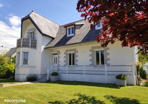 Une maison à deux étages, avec une façade blanche, un toit gris et un petit balcon à l’étage supérieur.