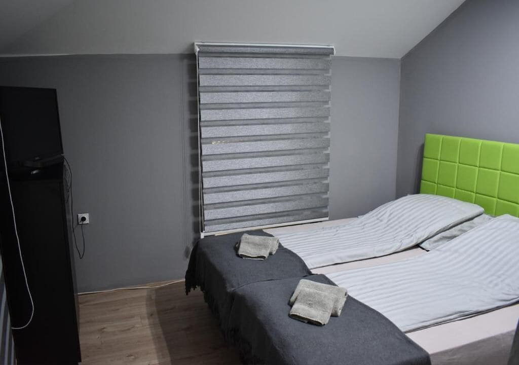 A bedroom with two beds, grey walls, a green headboard, and a window with blinds.