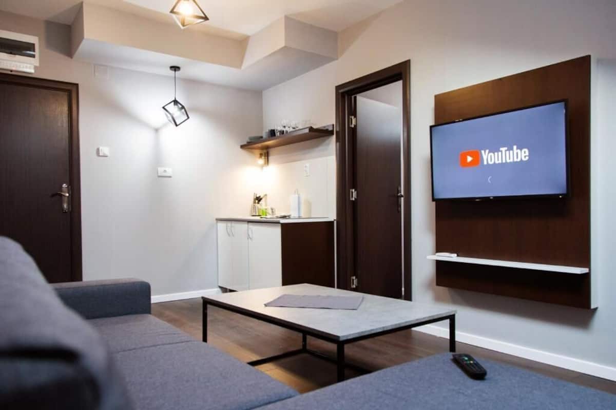 Un salon moderne avec un téléviseur à écran plat fixé au mur, un canapé, une table basse et un coin cuisine à l’arrière-plan.