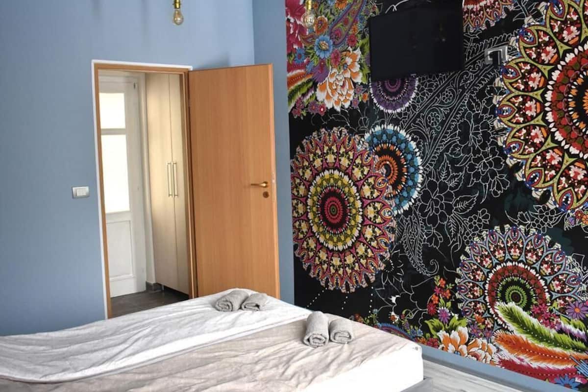 Une pièce avec un lit, une porte et une tapisserie colorée au mur.