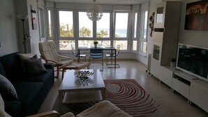 Una sala de estar moderna con un sofá, sillas, una mesa de centro y un televisor. Amplias ventanas ofrecen vistas a la playa y palmeras.