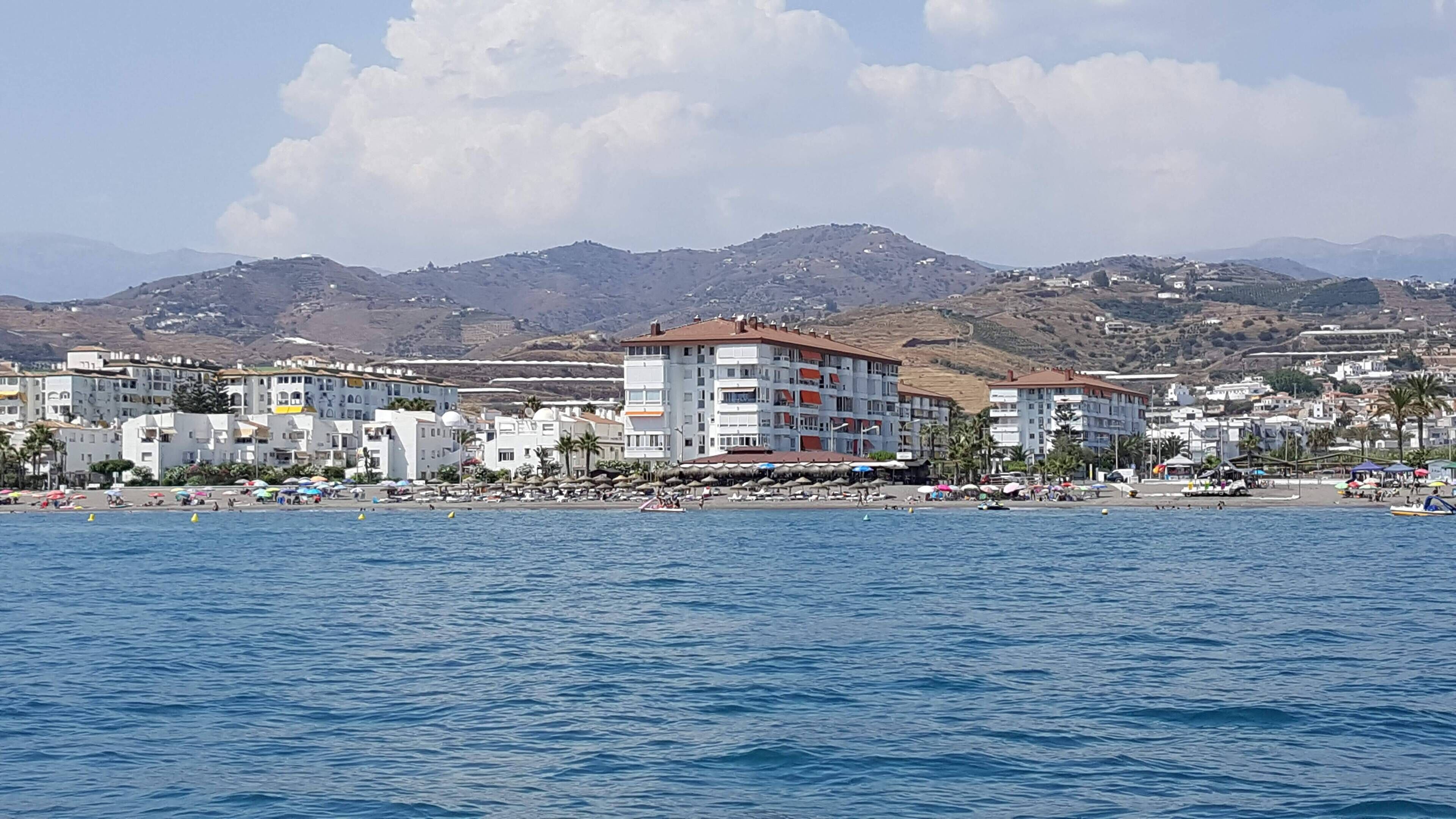 A coastal town with buildings along the waterfront, a beach with umbrellas, and mountains in the background.