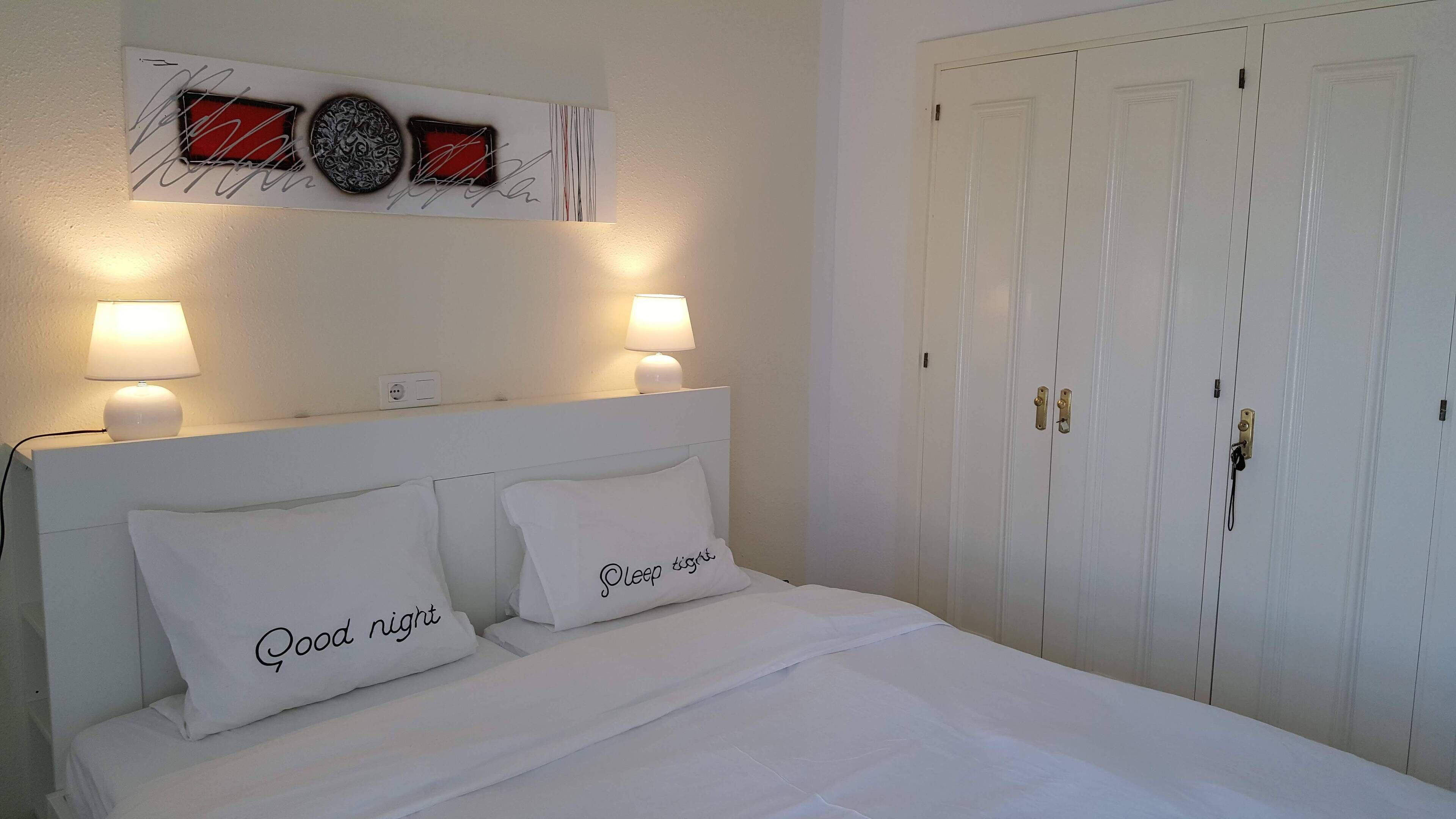 Een net opgemaakt bed met twee kussens, een aan de muur hangend kunstwerk en een witte garderobe met twee deuren.