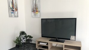 Een moderne woonkamer met een flatscreen televisie op een houten standaard, een potplant en twee abstracte schilderijen aan de muur.