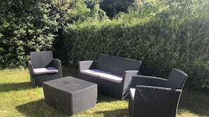 Een buitenmeubelset van riet met een tafel en stoelen, geplaatst op een gazon met weelderige begroeiing op de achtergrond.