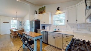 Una cocina moderna con armarios blancos, un refrigerador de acero inoxidable, una estufa a gas y un comedor con mesa y sillas de madera.