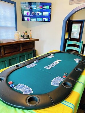 A pool table with cards and chips, a television displaying sports channels, and a wooden desk with a pitcher.
