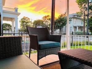 En fletstol med pude på en balkon med gelænder, udsigt til en hvid bygning og en solnedgang i baggrunden.