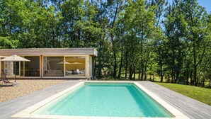 Ein moderner Poolbereich im Freien mit Holzterrasse, einer gläsernen Lounge mit Sitzgelegenheiten und einer Kulisse aus dichtem Baumbestand.