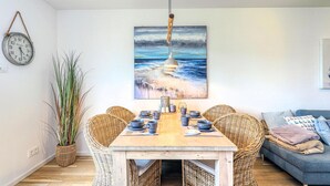 Un comedor con una mesa de madera dispuesta para cuatro personas, sillas de mimbre, un cuadro de una playa y un reloj colgante.