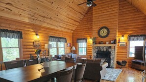 A wooden cabin interior with a dining area, living room, fireplace, and a clock on the wall.