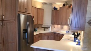 A kitchen with wooden cabinets, a stainless steel refrigerator, a white countertop, and two stools.
