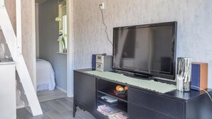 Una habitación con un televisor de pantalla grande sobre un soporte negro, una mesita con un jarrón de flores y una ventana con vistas a un balcón.