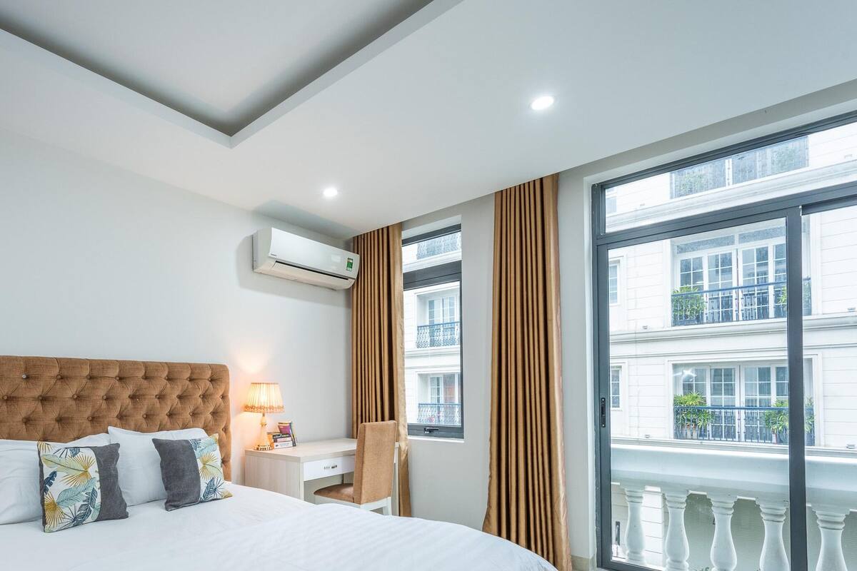 Une chambre d’hôtel avec une tête de lit capitonnée, un lit avec du linge de lit blanc, deux oreillers, une table de chevet avec une lampe, une fenêtre avec des rideaux et un climatiseur fixé au mur.