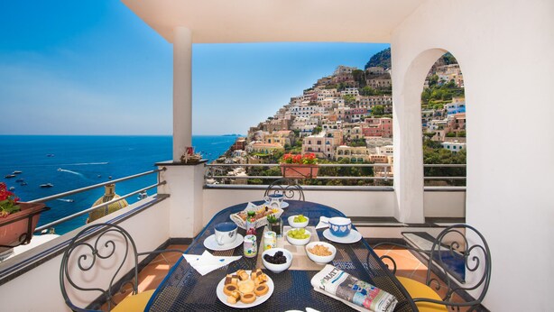Un balcon avec une table préparée pour le petit-déjeuner, offrant une vue sur une ville côtière et la mer.