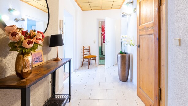 A hallway with a wooden door, a large vase with flowers, a chair, and a table with a lamp.