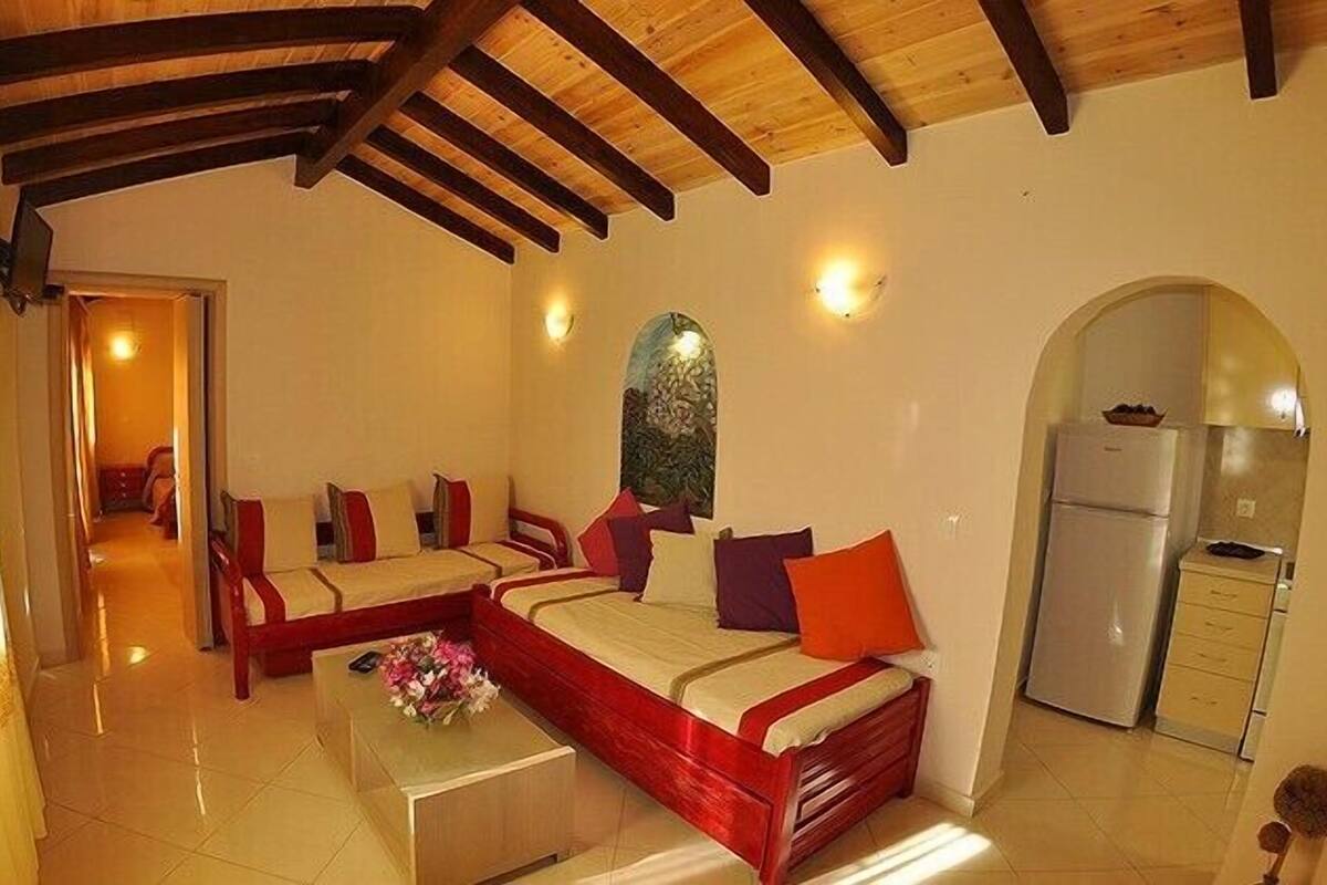 A bedroom with a wooden ceiling, a bed with red and beige bedding, a small table with a vase, and a kitchen area with a refrigerator.