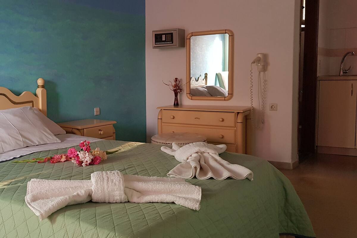 A neatly made bed with white linens, a green quilt, and a bouquet of flowers. There is a wooden nightstand with a lamp, a dresser with a mirror, and a telephone.