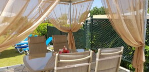 An outdoor seating area with wicker chairs and a glass table, covered by a canopy with sheer curtains. - Elena's Seaside Holiday House (Corfu)