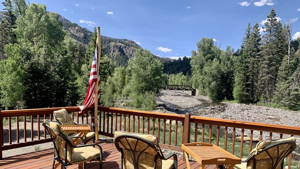Een terras met buitenmeubilair en een Amerikaanse vlag, met uitzicht op een rivier en beboste bergen.