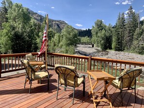 A deck with outdoor furniture and an American flag, overlooking a river and forested mountains.