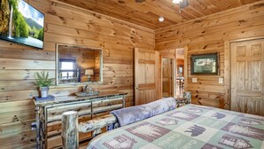 A wooden cabin room with a bed, a dresser, a mirror, and a television on the wall. - Panoramic View - Suite 2: Brand New Hot Tub - Room w/ French Doors to Porch (sevierville)