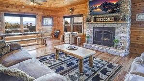 A cozy log cabin interior with a stone fireplace, wooden furniture, and a scenic view through large windows. - Panoramic View - Suite 2: Brand New Hot Tub - Room w/ French Doors to Porch (sevierville)
