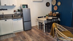 Une cuisine compacte avec des meubles blancs, un four à micro-ondes encastré et un réfrigérateur. On y trouve un coin repas avec une table et des chaises, un mur bleu orné d’assiettes décoratives et un fauteuil en osier.