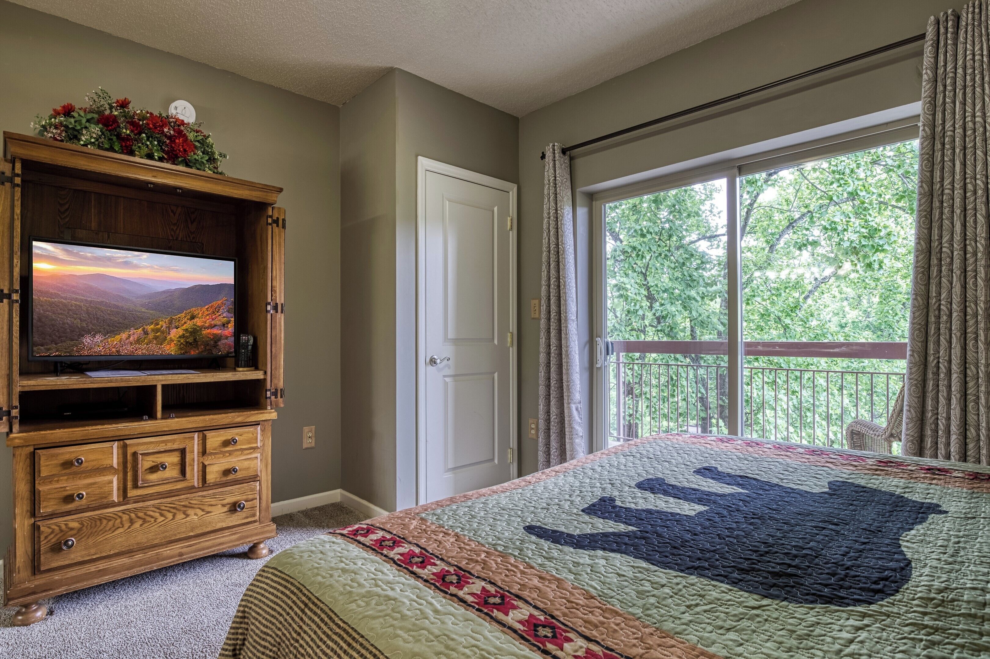 A bedroom with a large bed, a wooden dresser with a TV, a window with curtains, and a balcony.